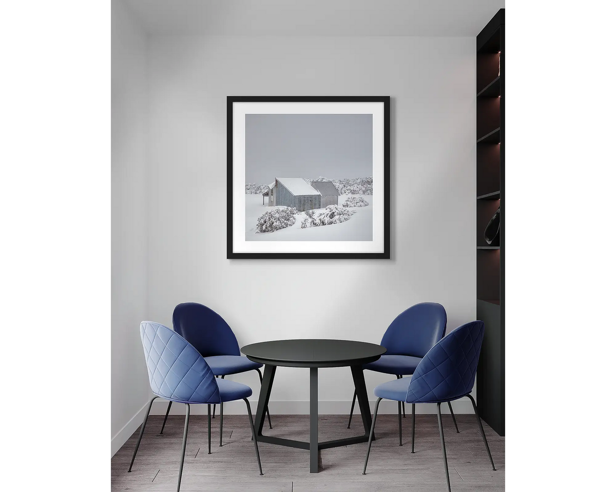 Blowhard. Framed, Alpine hut in snow, artwork with a black frame, hung above a meeting room table.