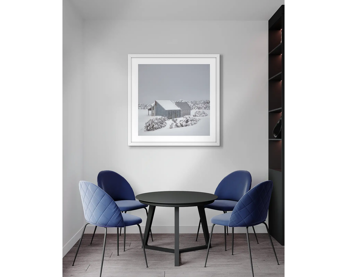 Blowhard. Framed, Alpine hut in snow, artwork with a white frame, hung above a meeting room table.