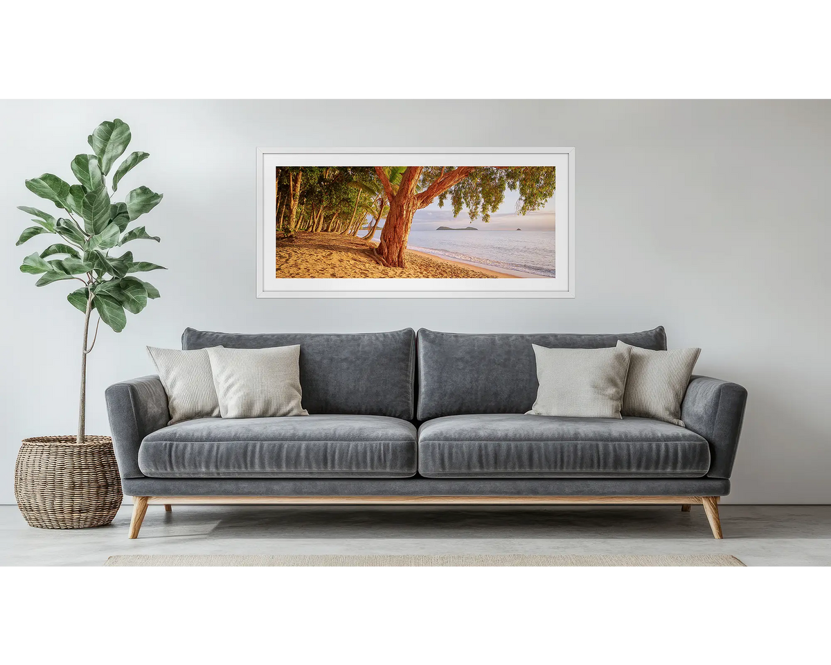 Cairns Escape. Framed wall art print of a sunrise on a tropical beach, with a white frame, hung above a couch.