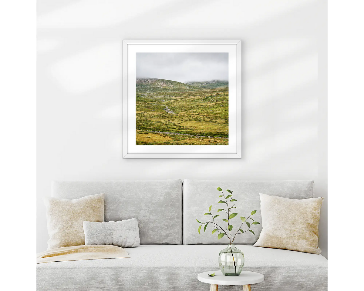 Drizzle. Snowy Mountains wall art print with white frame hung above couch.