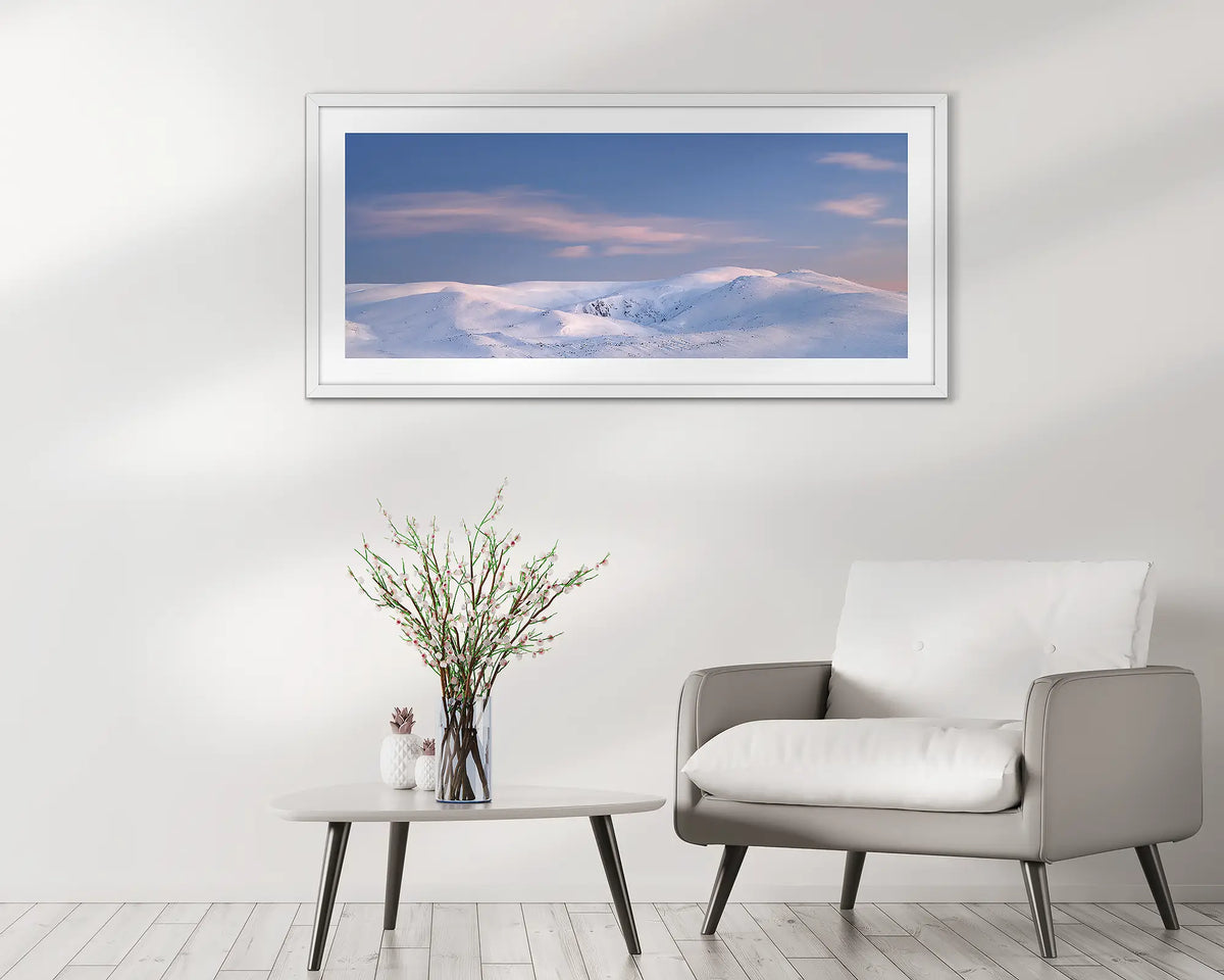 Early Morning. Snowy Mountains sunrise wall art print, with a white frame, hung above a chair.