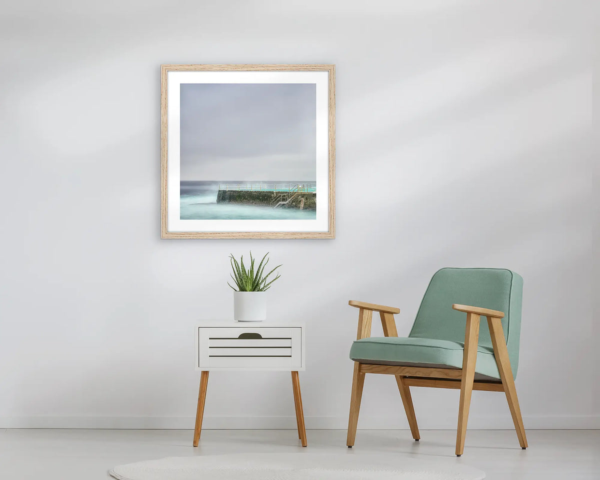 Moody Bondi. Framed wall art print, in an Oak frame, of Icebergs at Bondi beach, hanging above a table. 