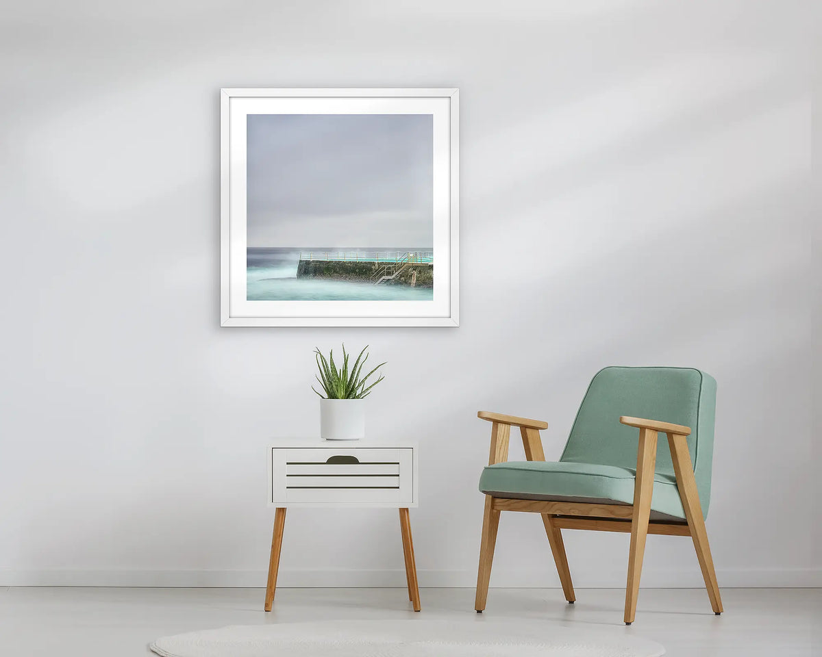 Moody Bondi. Framed wall art print, in a white frame, of Icebergs at Bondi beach, hanging above a table. 